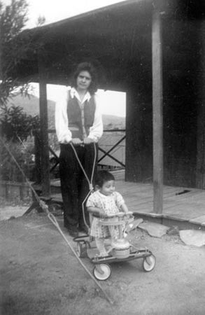 A black and white photo of a woman pushing a child in a small cart in front of a wooden porch, with hills in the background.