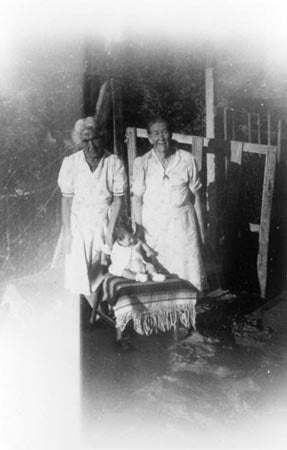 Black-and-white photo of two elderly women and a child sitting between them on a stool.