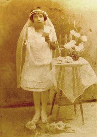 A young girl wearing a veil, dressed in a light colored dress holds a book and a candle next to a lace-covered table with flowers.