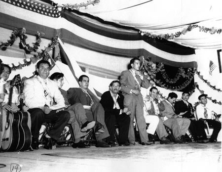 A group of men in suits on a decorated stage, one speaking into a microphone.