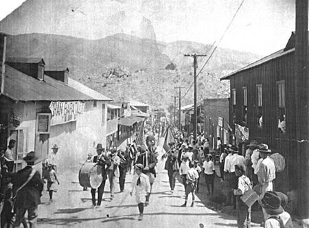 A black and white photograph of a parade with a band marching down a street lined with buildings and onlookers.