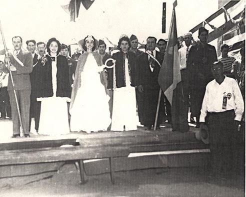 Black and white photo of a group of people on a stage during a ceremony, with women in gowns and a man holding a flag.