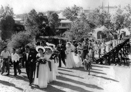 A vintage photo of a wedding party from the early 20th century, with a bride, groom, and guests walking near a wooden bridge.