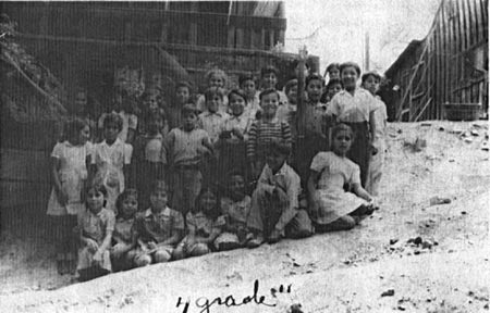 A black and white photo of a group of children posing on a slope, with wooden structures in the background. Written at the bottom is '4 grade'.