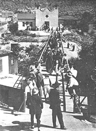 People walking on a wooden bridge towards a church in the background.