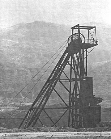 A black and white photo of a mining headframe with mountains in the background.