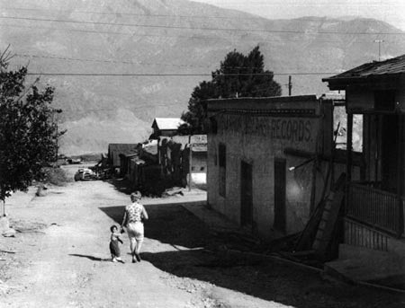 A dusty street in a small village with mountains in the background, featuring a woman and child walking, and buildings on the right.