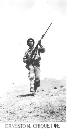 A black and white photo of a soldier in a historical military uniform carrying a rifle, walking on a barren hill.