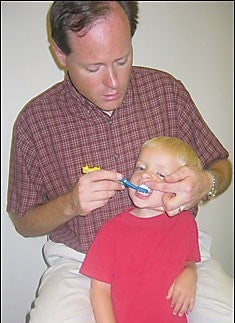 Picture of caregiver that is properly brushing the teeth of a child. Caregiver is behind the child and brushing the teeth of the child that is turned slightly sideways in the caregiver's lap.
