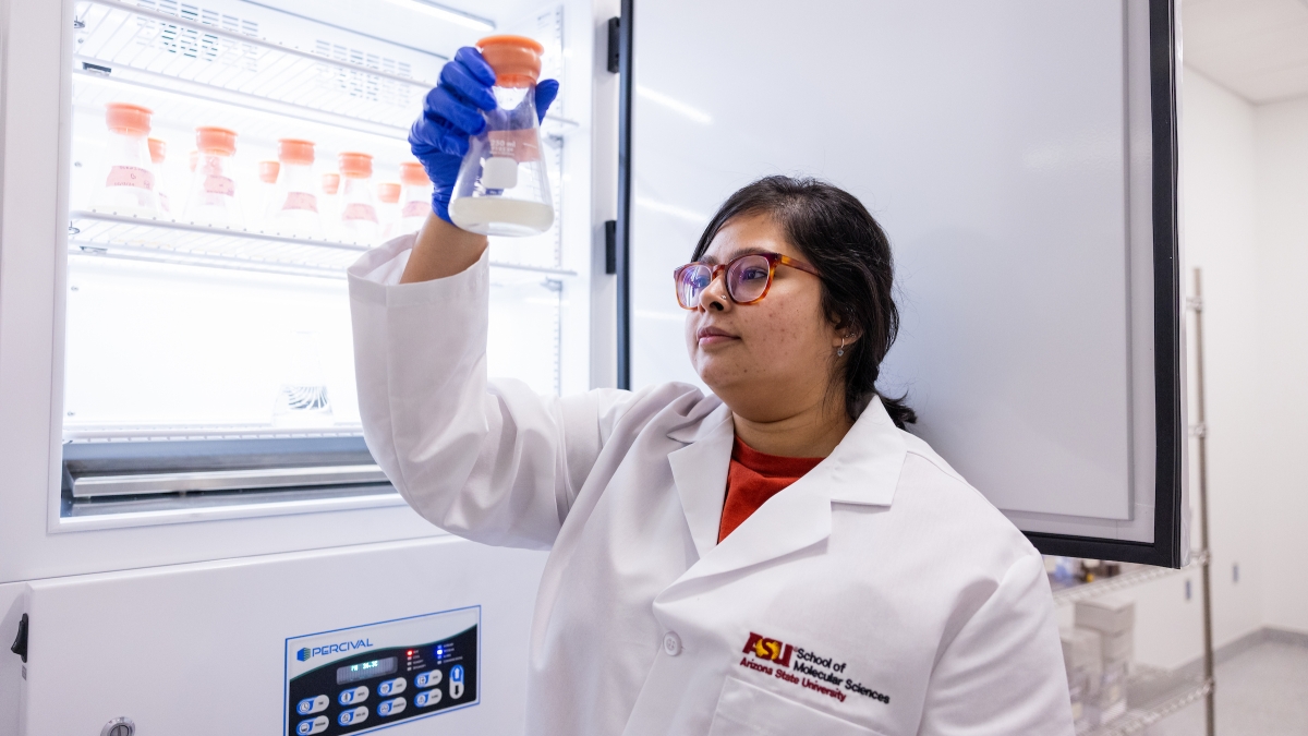 A young woman wearing glasses, blue gloves and a white lab coat holds out a beaker full of liquid in a lab